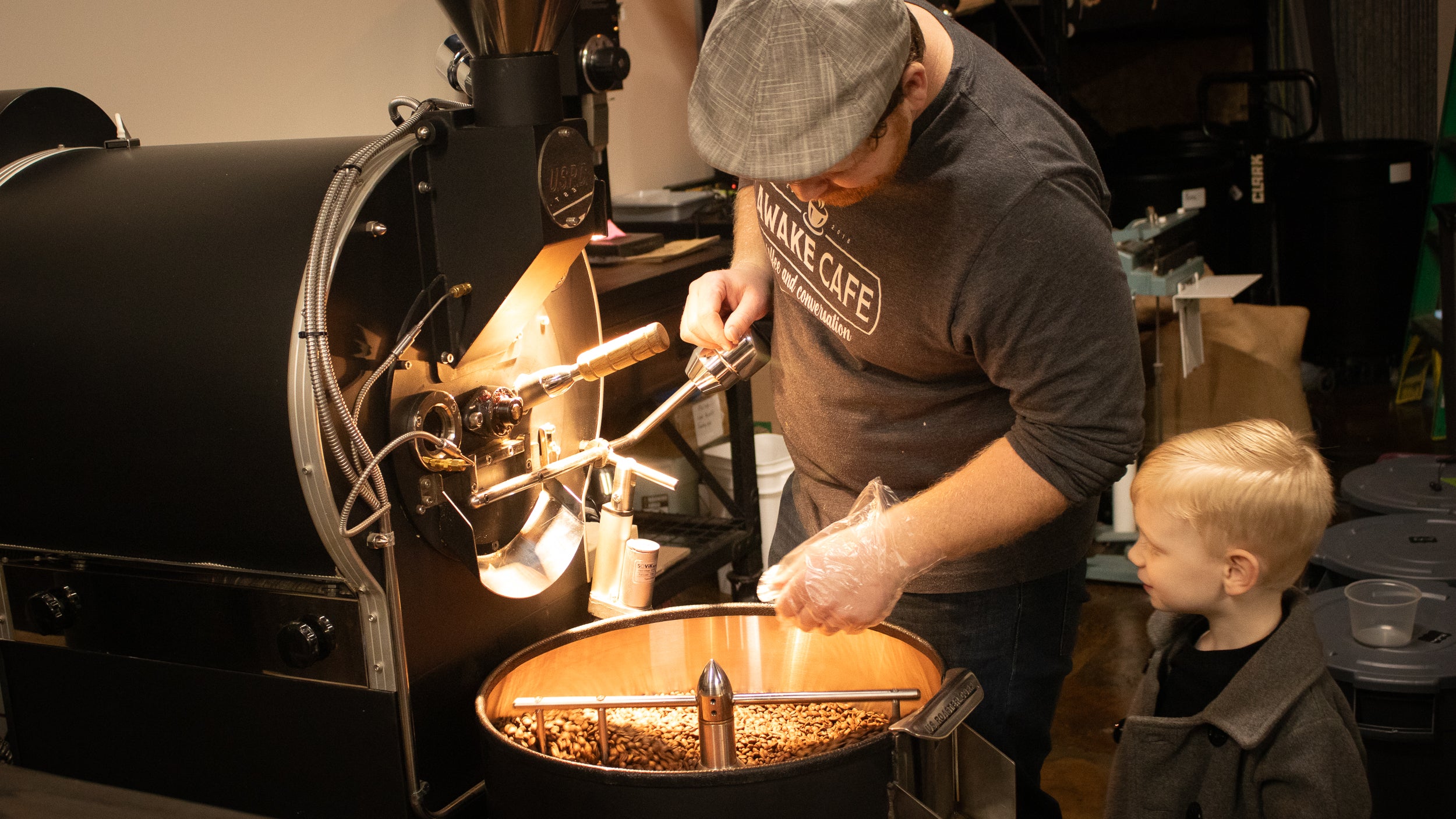 Person operating a coffee roasting machine with a child observing.